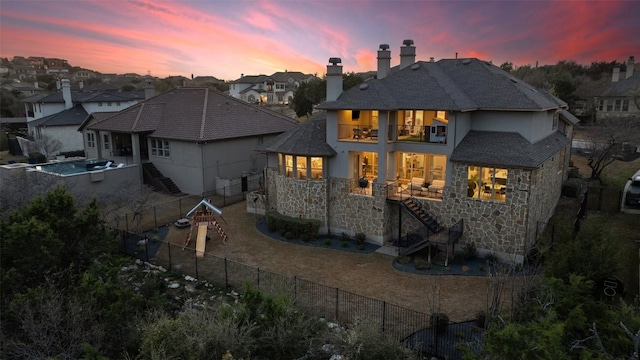 back of house at dusk with stone siding, a balcony, stairs, and a fenced backyard