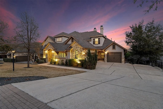 french provincial home featuring stone siding, an attached garage, concrete driveway, and a chimney