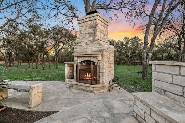 patio terrace at dusk featuring a yard, an outdoor stone fireplace, and fence