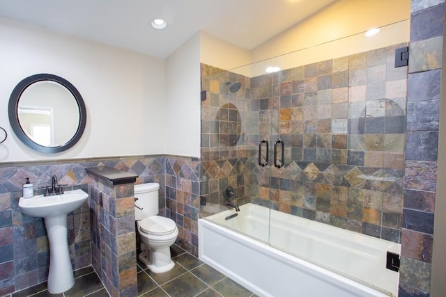 bathroom with a wainscoted wall, tile patterned flooring, tile walls, toilet, and combined bath / shower with glass door