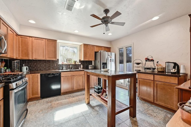 kitchen with brown cabinetry, a ceiling fan, stainless steel appliances, decorative backsplash, and dark countertops