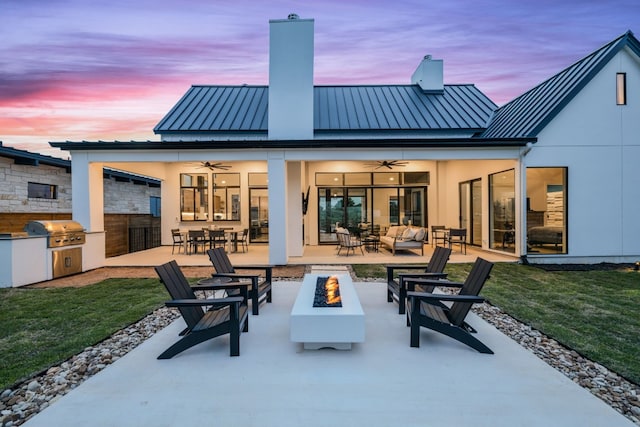 back of house at dusk featuring a standing seam roof, an outdoor living space with a fire pit, metal roof, an outdoor kitchen, and a chimney