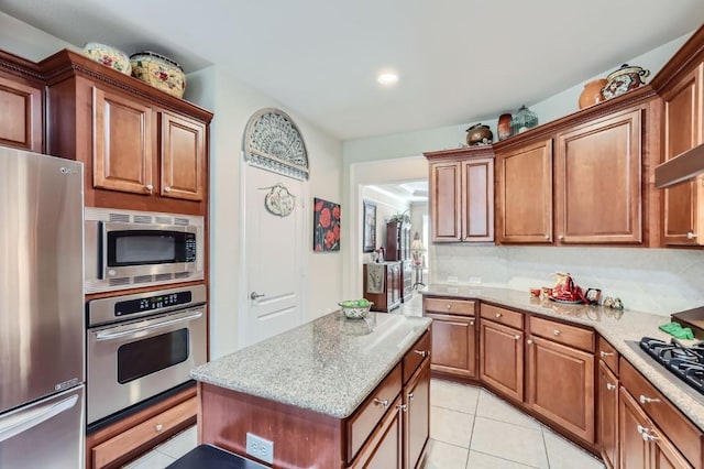 kitchen with a center island, appliances with stainless steel finishes, light tile patterned flooring, brown cabinetry, and light stone countertops