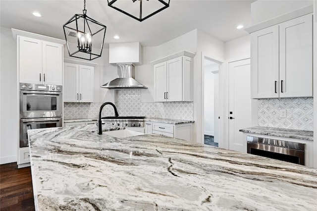 kitchen with light stone countertops, stainless steel double oven, dark wood-style flooring, white cabinetry, and wall chimney exhaust hood