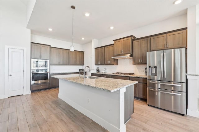 kitchen with a sink, under cabinet range hood, stainless steel appliances, light wood finished floors, and light stone countertops