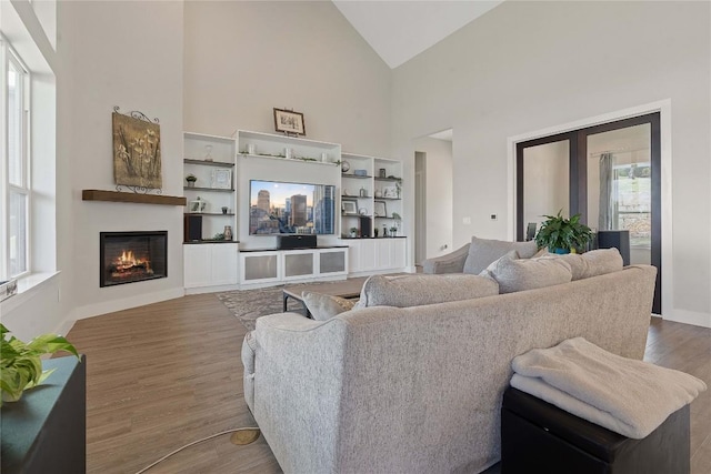 living room with a warm lit fireplace, high vaulted ceiling, baseboards, and wood finished floors