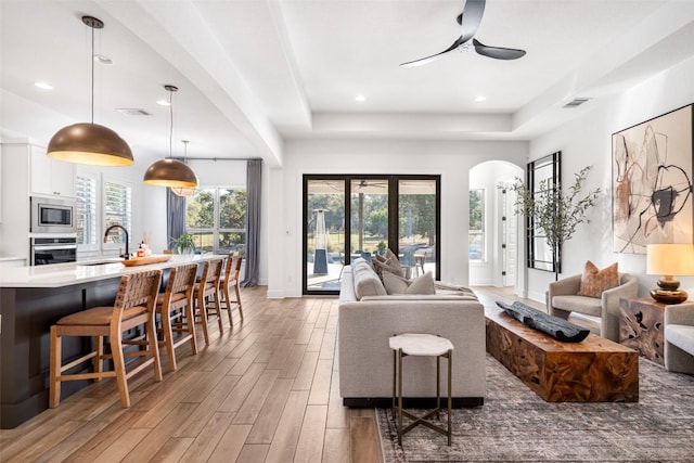 living area featuring visible vents, light wood-style floors, arched walkways, a raised ceiling, and a ceiling fan