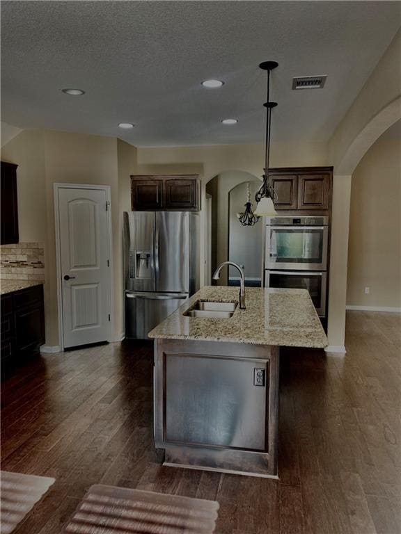 kitchen featuring visible vents, a sink, appliances with stainless steel finishes, arched walkways, and a kitchen island with sink