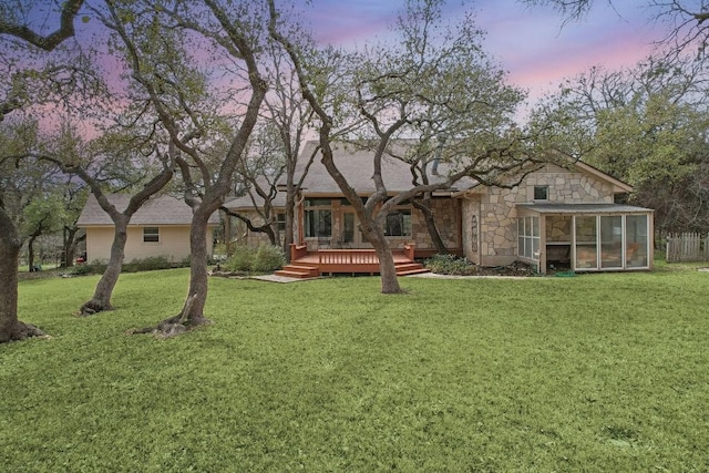 back of house at dusk featuring a deck, a yard, stone siding, and a sunroom