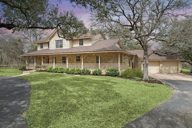 farmhouse-style home featuring driveway, covered porch, a front lawn, a garage, and stone siding