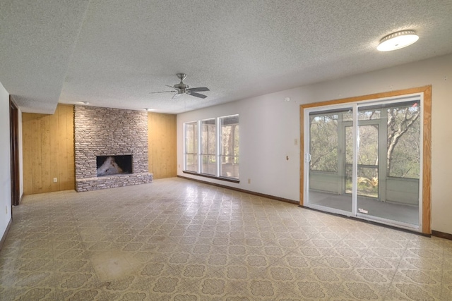 unfurnished living room with wooden walls, a ceiling fan, baseboards, a stone fireplace, and a textured ceiling