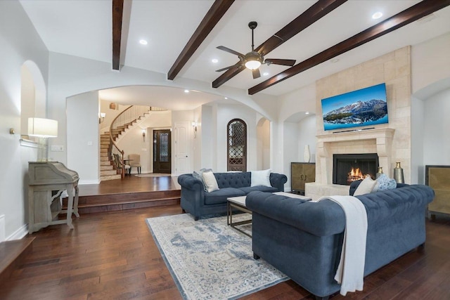 living room featuring ceiling fan, stairs, beam ceiling, a fireplace, and dark wood-style floors