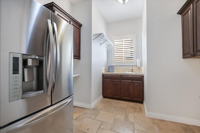 kitchen featuring stone tile flooring, stainless steel fridge, dark brown cabinetry, light countertops, and baseboards