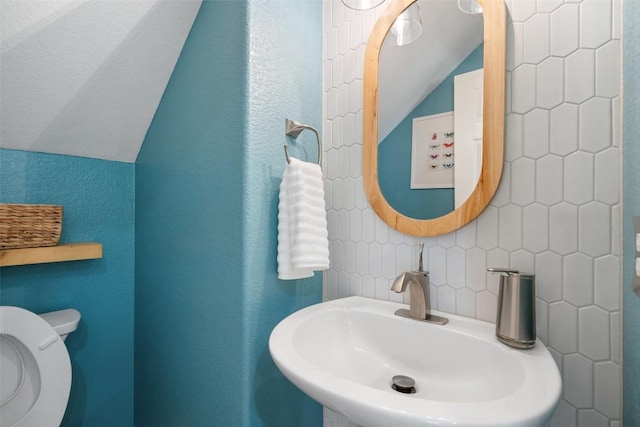 half bath with a sink, vaulted ceiling, toilet, a textured wall, and backsplash