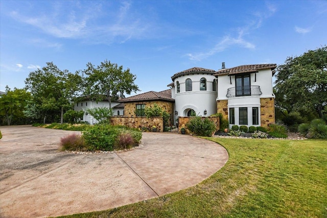 mediterranean / spanish home featuring stucco siding, a front lawn, stone siding, a balcony, and a tiled roof
