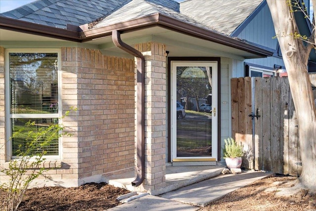 doorway to property featuring a shingled roof, brick siding, and fence