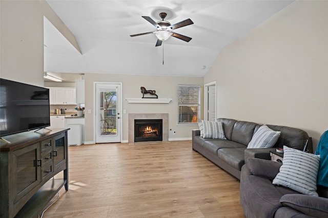 living room with ceiling fan, vaulted ceiling, a tile fireplace, baseboards, and light wood-style flooring