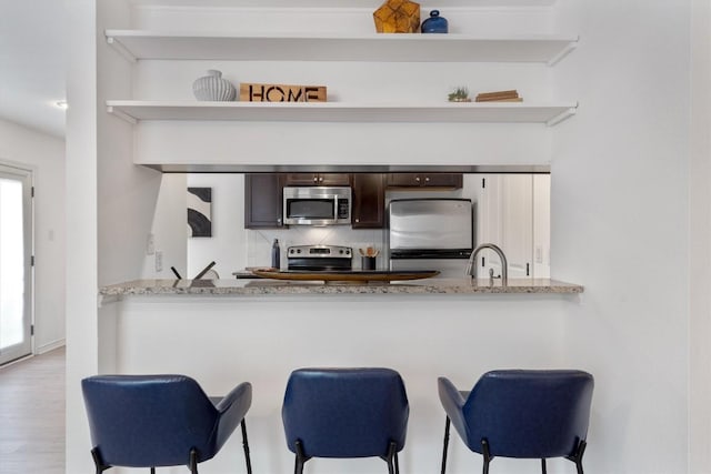 kitchen with appliances with stainless steel finishes, dark brown cabinetry, backsplash, open shelves, and a breakfast bar