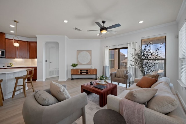 living area with visible vents, light wood-type flooring, ornamental molding, arched walkways, and a ceiling fan