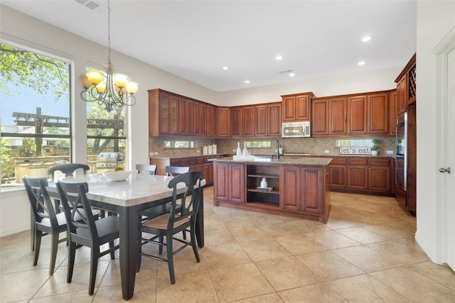 kitchen featuring stainless steel appliances, backsplash, a chandelier, light stone counters, and a center island with sink