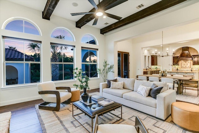 living area featuring visible vents, beamed ceiling, ceiling fan with notable chandelier, hardwood / wood-style flooring, and baseboards