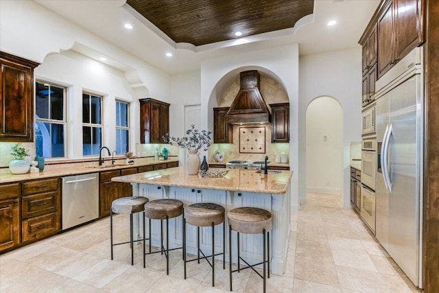 kitchen featuring premium range hood, built in appliances, an island with sink, a tray ceiling, and a sink