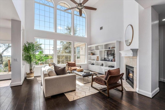 living area featuring a ceiling fan, a towering ceiling, a tiled fireplace, and wood-type flooring