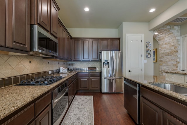 kitchen with dark brown cabinetry, dark wood-style floors, light stone counters, arched walkways, and appliances with stainless steel finishes