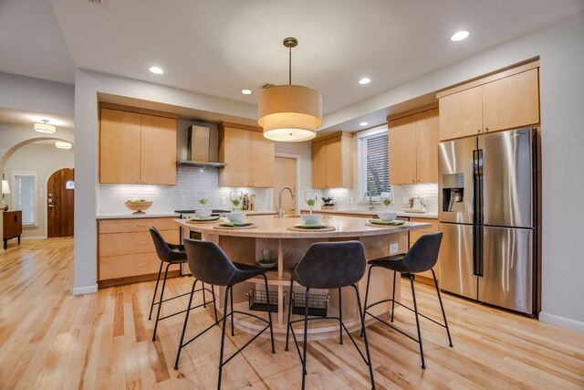 kitchen with wall chimney range hood, a breakfast bar area, light countertops, stainless steel fridge with ice dispenser, and light brown cabinets