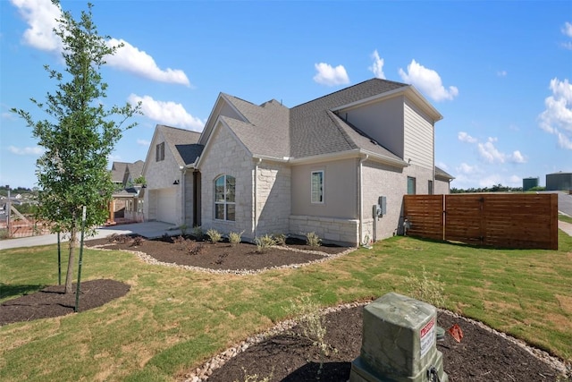 view of side of home with driveway, stone siding, fence, a lawn, and a shingled roof