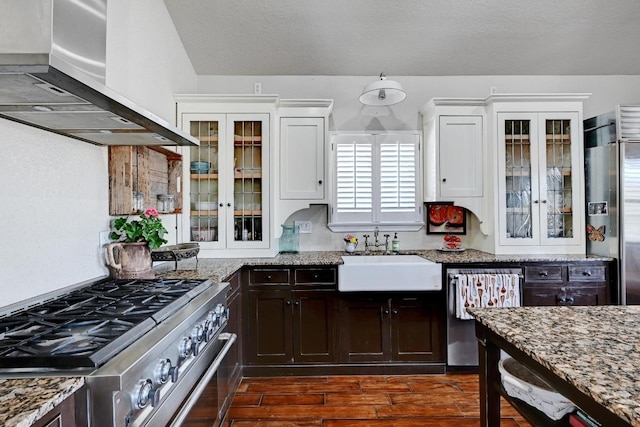 kitchen with a sink, appliances with stainless steel finishes, glass insert cabinets, wall chimney range hood, and dark brown cabinets