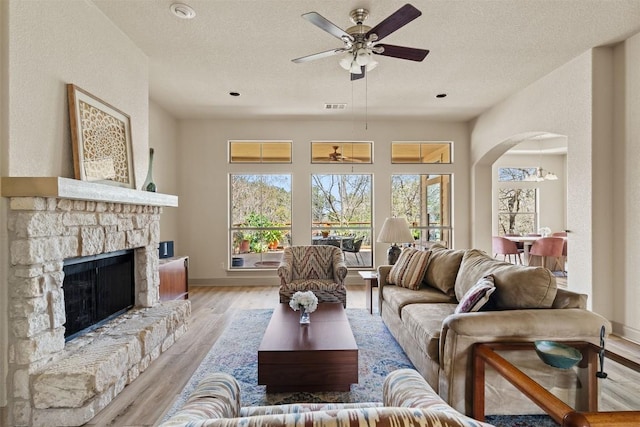 living room with light wood finished floors, a stone fireplace, visible vents, arched walkways, and ceiling fan