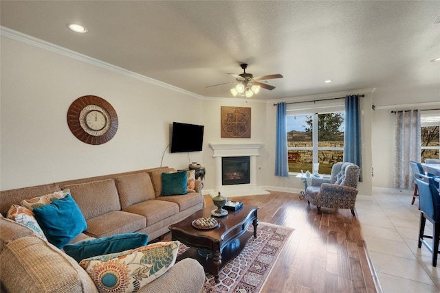 living room featuring a glass covered fireplace, light wood-style floors, ceiling fan, baseboards, and ornamental molding