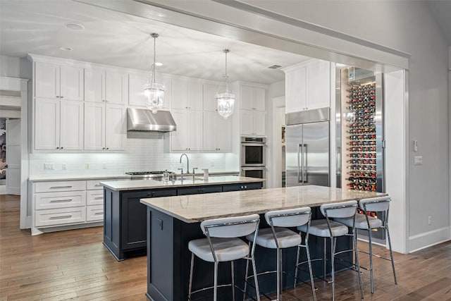 4229 Verano Dr, Austin, TX 78735 - kitchen featuring stainless steel appliances, a center island with sink, under cabinet range hood, tasteful backsplash, and white cabinets