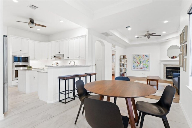 dining space with ceiling fan, a raised ceiling, recessed lighting, a glass covered fireplace, and baseboards