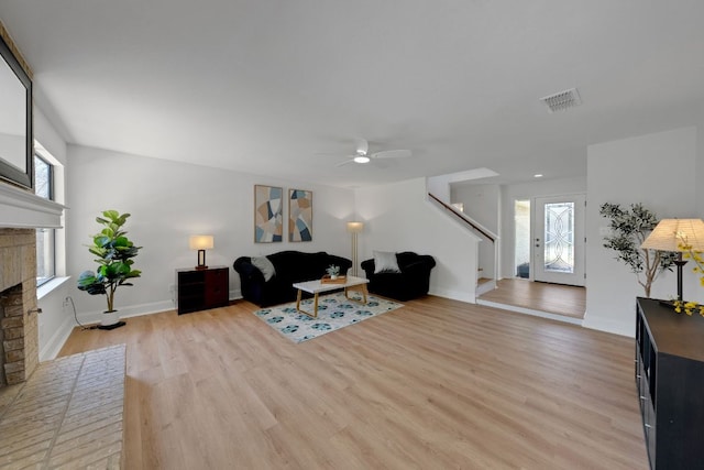 living room with a brick fireplace, stairs, light wood-type flooring, visible vents, and a ceiling fan
