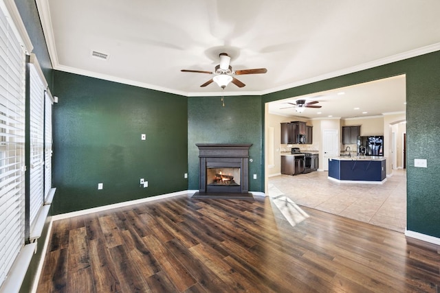 unfurnished living room with crown molding, visible vents, a warm lit fireplace, ceiling fan, and wood finished floors
