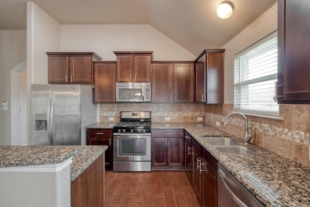 kitchen featuring stainless steel appliances, a sink, vaulted ceiling, light stone counters, and decorative backsplash