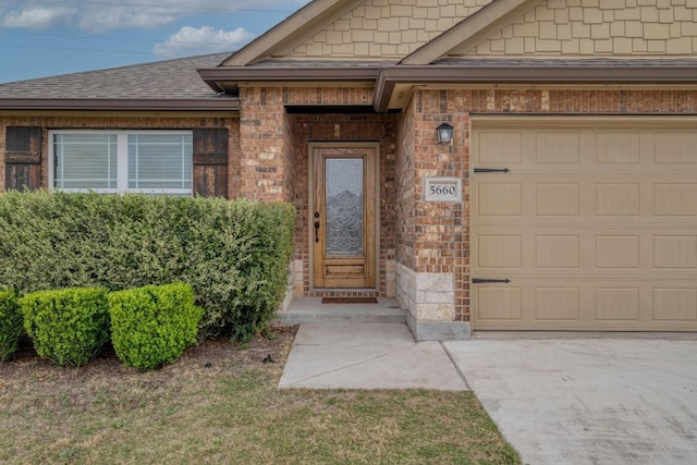 entrance to property with brick siding, roof with shingles, an attached garage, and driveway