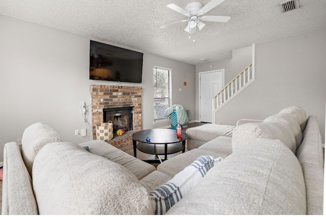 living room featuring a brick fireplace, stairs, a textured ceiling, visible vents, and a ceiling fan