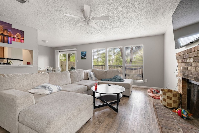 living room with wood finished floors, a textured ceiling, a brick fireplace, baseboards, and ceiling fan