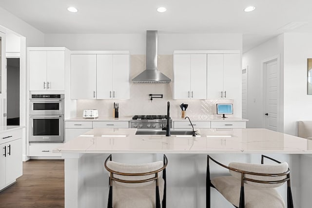 kitchen featuring double oven, wall chimney exhaust hood, dark wood-style flooring, and a breakfast bar