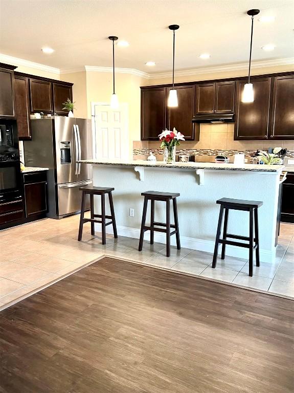 kitchen with stainless steel fridge with ice dispenser, black microwave, tasteful backsplash, under cabinet range hood, and a breakfast bar area