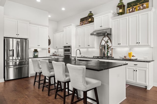 kitchen featuring a sink, dark countertops, stainless steel appliances, under cabinet range hood, and a center island with sink