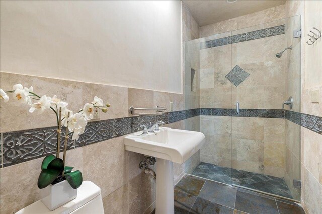 bathroom featuring a stall shower, toilet, a wainscoted wall, tile walls, and a sink