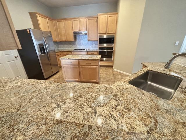 kitchen with light stone counters, a sink, appliances with stainless steel finishes, under cabinet range hood, and decorative backsplash