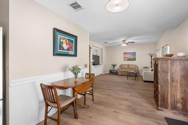 dining area with light wood-style flooring, a wainscoted wall, visible vents, a ceiling fan, and a textured ceiling