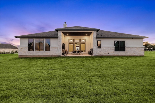 back of house at dusk with a patio, stucco siding, a shingled roof, and a yard