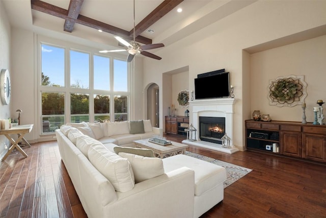 living area with dark wood-style flooring, a lit fireplace, a towering ceiling, arched walkways, and beamed ceiling