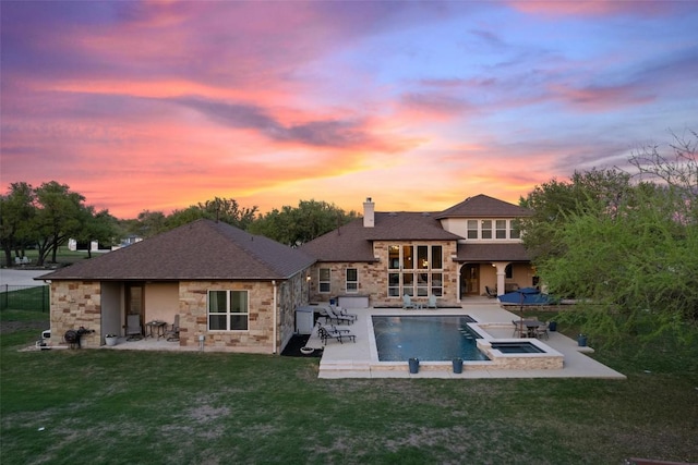back of property at dusk with a patio area, stone siding, roof with shingles, a chimney, and a yard